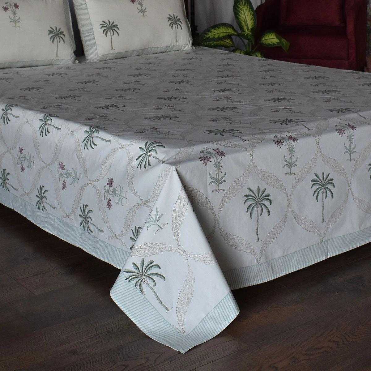A bed dressed with a white tablecloth featuring a repeating palm tree and floral pattern, with matching pillows visible at the head. The room setting includes a dark wood floor and a red upholstered chair in the background.