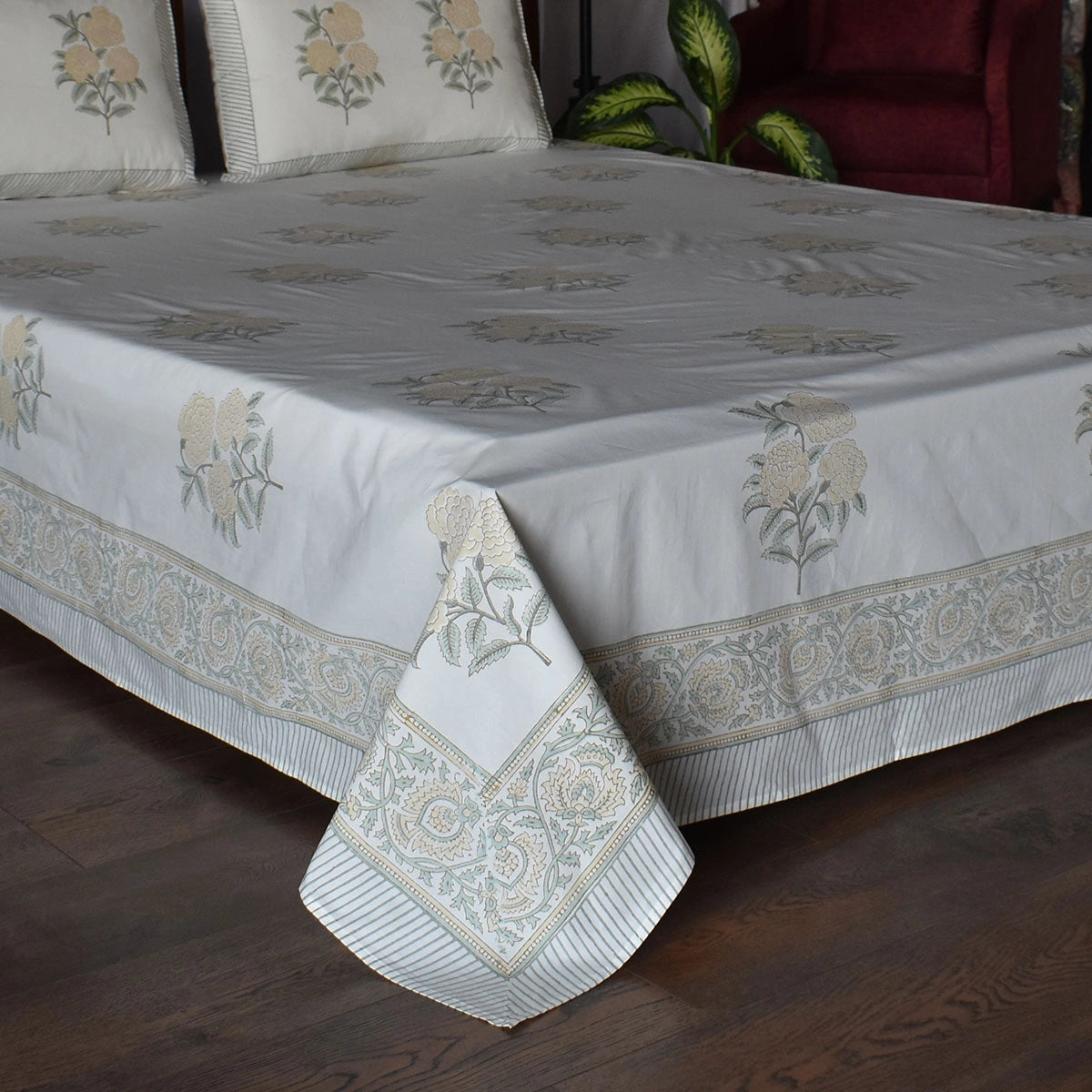 A white cotton bed sheet with a beige and green floral pattern and a decorative border, draped over a bed in a bedroom setting with a dark wood floor.
