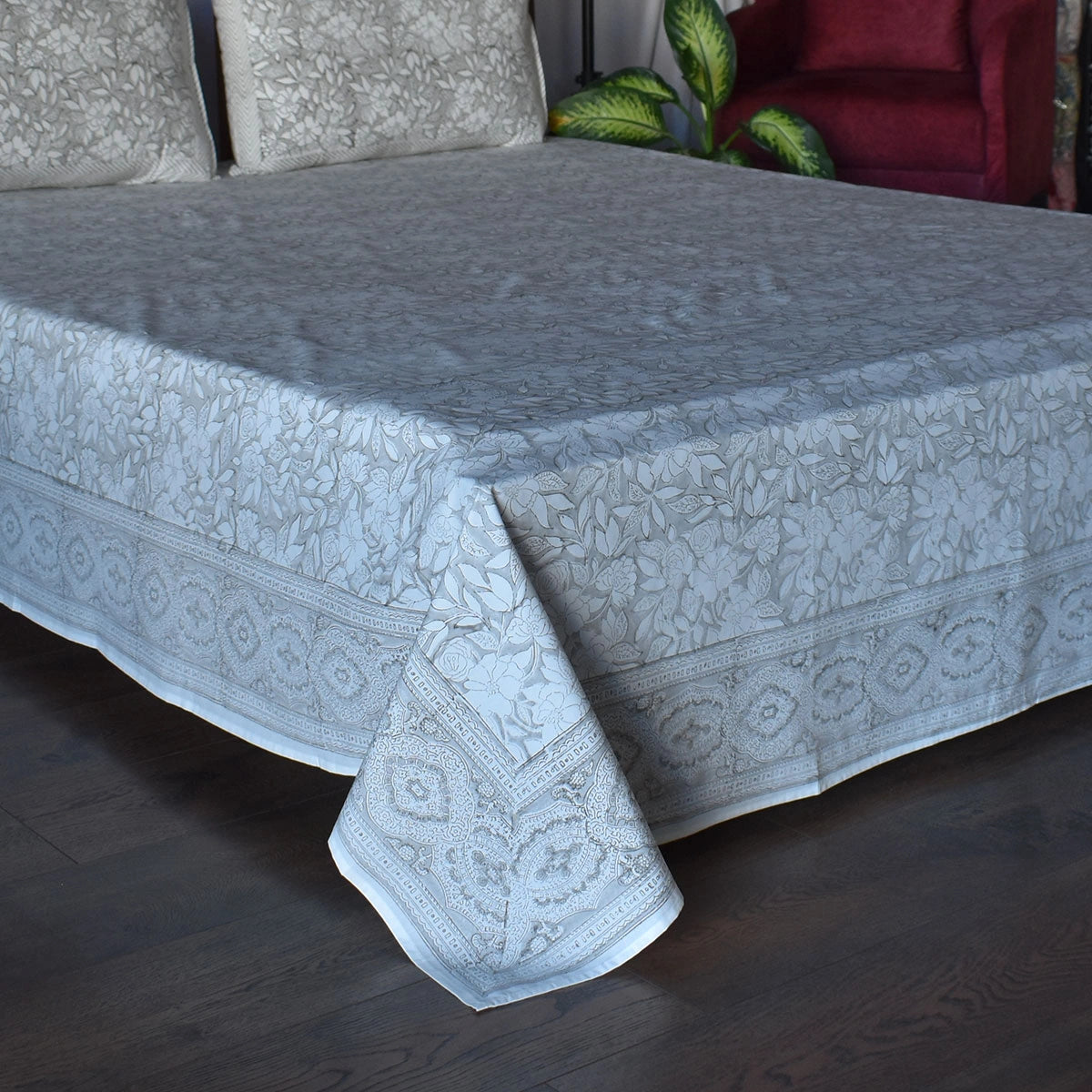 A light blue patterned cotton bed sheet with a matching pillowcase, draped over a bed on a dark wood floor with a red armchair and green plant in the background.