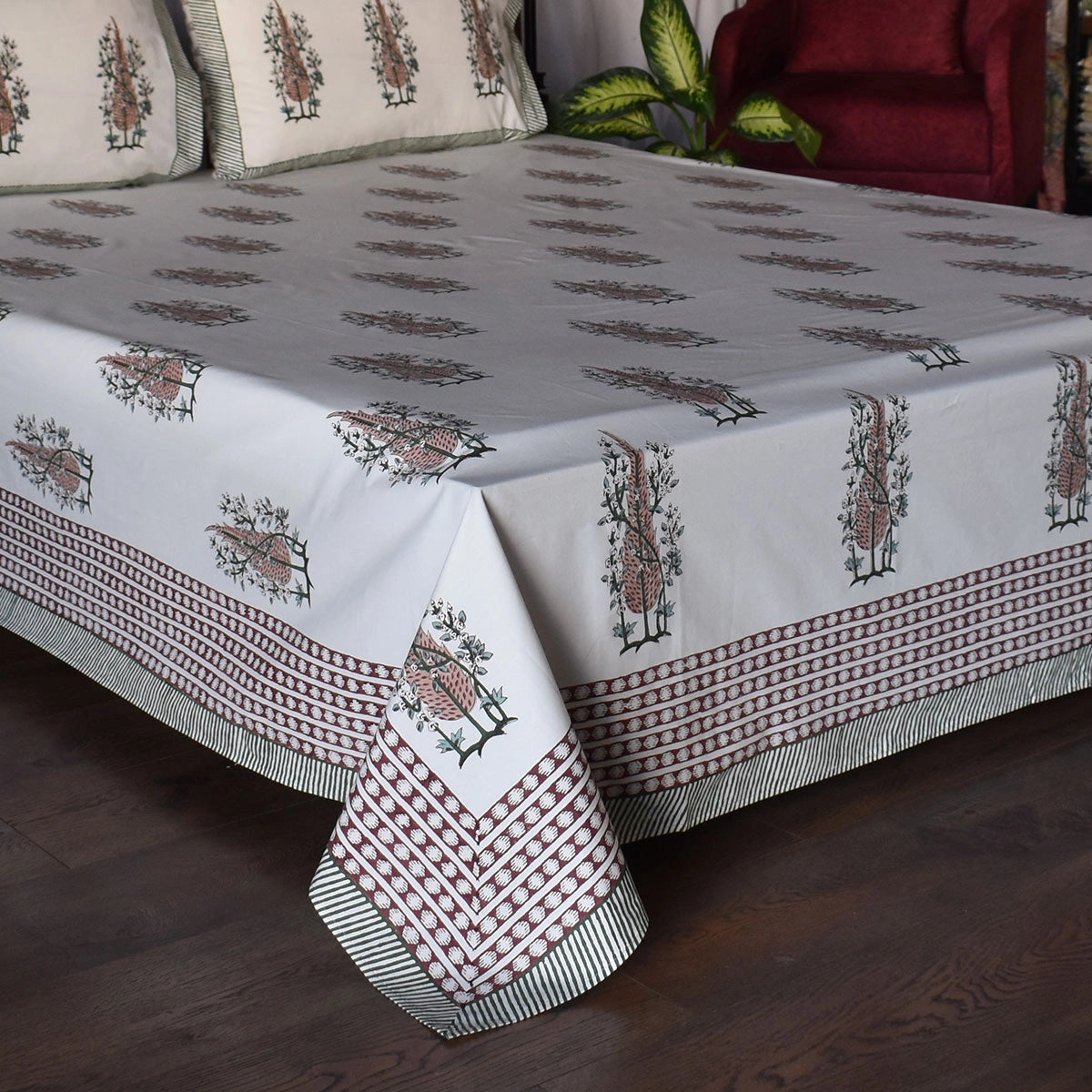 A white cotton bedspread with traditional block-printed floral motifs and striped borders, styled on a bed in a bedroom setting with matching pillows, a wooden floor, and a red armchair in the background.