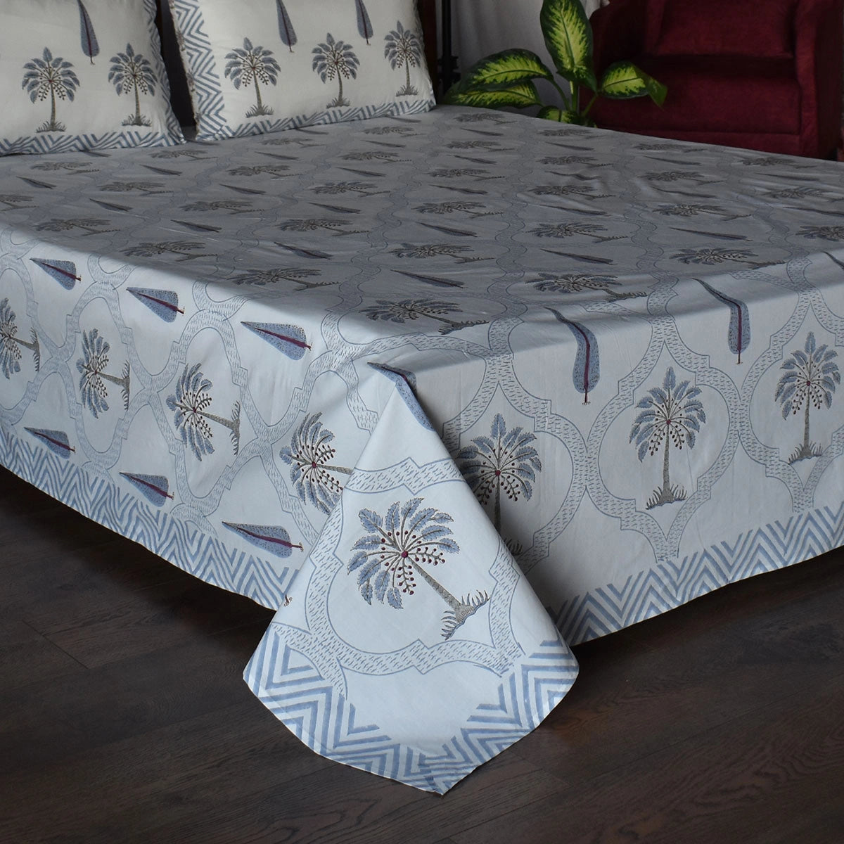 A bed dressed in a light blue and gray patterned bedspread or sheet set with matching pillowcases, featuring palm tree motifs and geometric border designs, shown in a bedroom setting with dark wood flooring and a red armchair in the background.