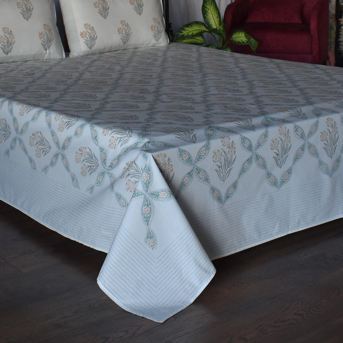 A light blue patterned bedsheet draped over a bed, featuring a repeating floral and geometric design in muted blue, beige, and white. The bed is set in a bedroom with dark wood flooring, matching patterned pillows, and a red armchair in the background.