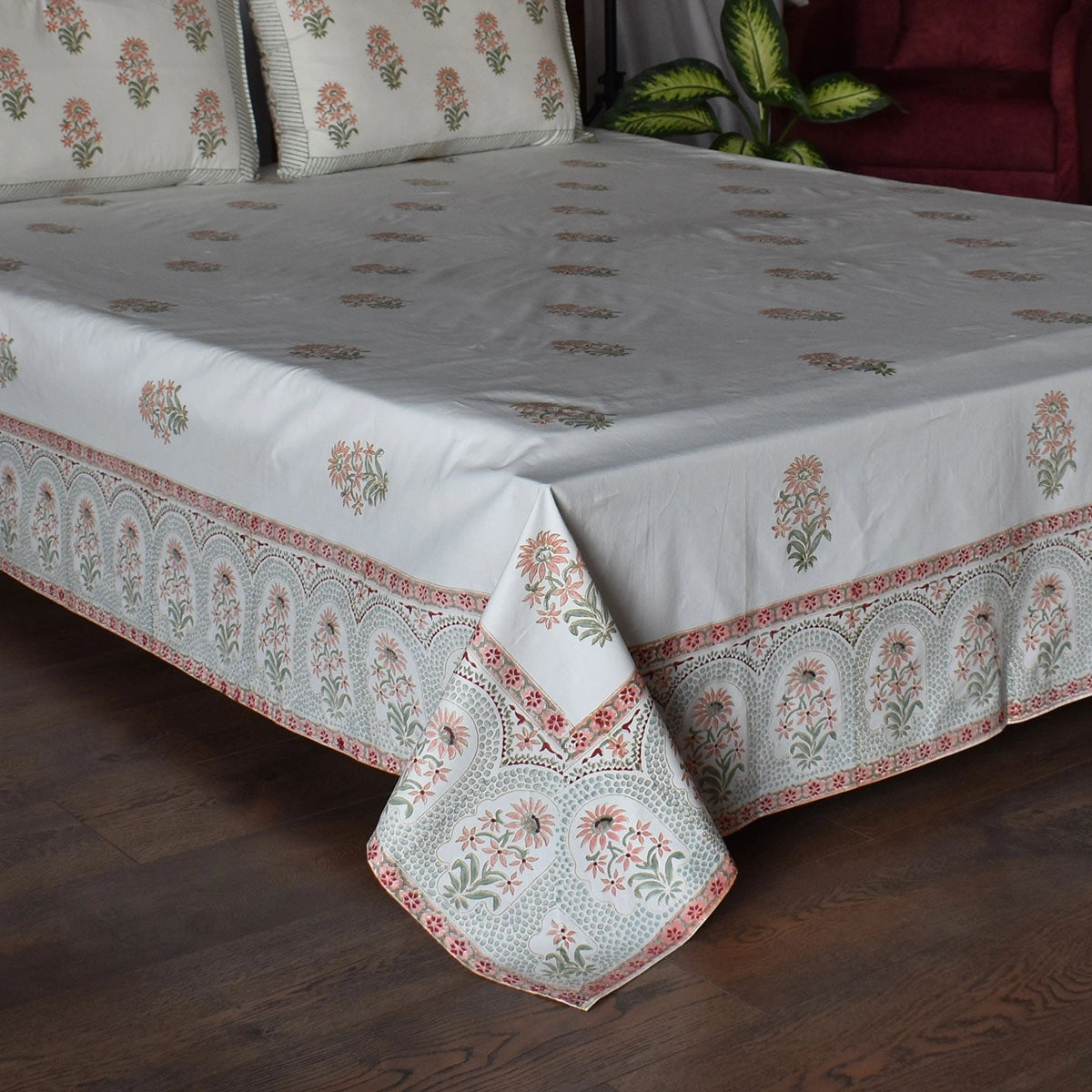 A light gray cotton bed sheet with intricate floral block prints and a detailed border design, draped over a bed in a bedroom setting with a dark wood floor.