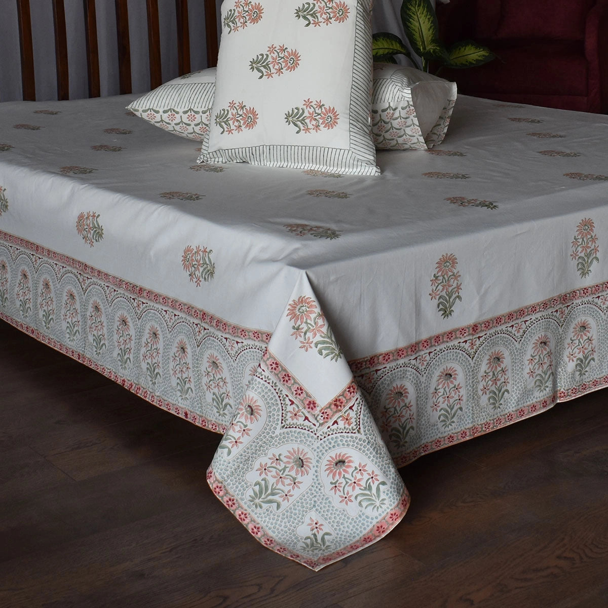A white cotton bed sheet with a detailed red and green floral border and matching printed pillows, styled on a bed in a bedroom setting.