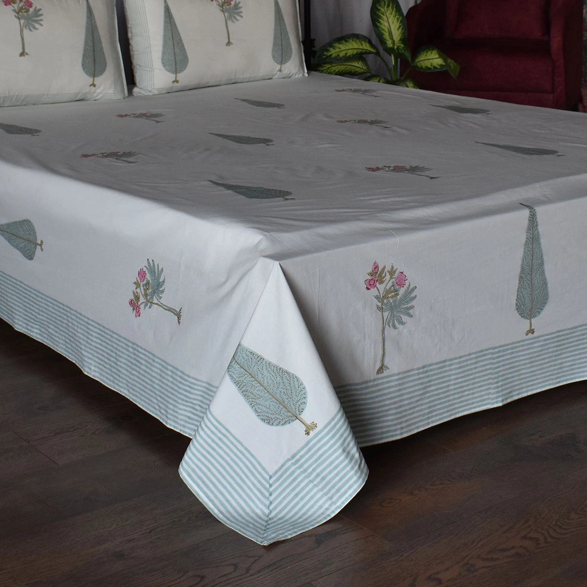 A white bedspread with a light blue striped border and printed botanical motifs, shown on a bed in a bedroom setting with matching pillows, a plant, and a red armchair visible.