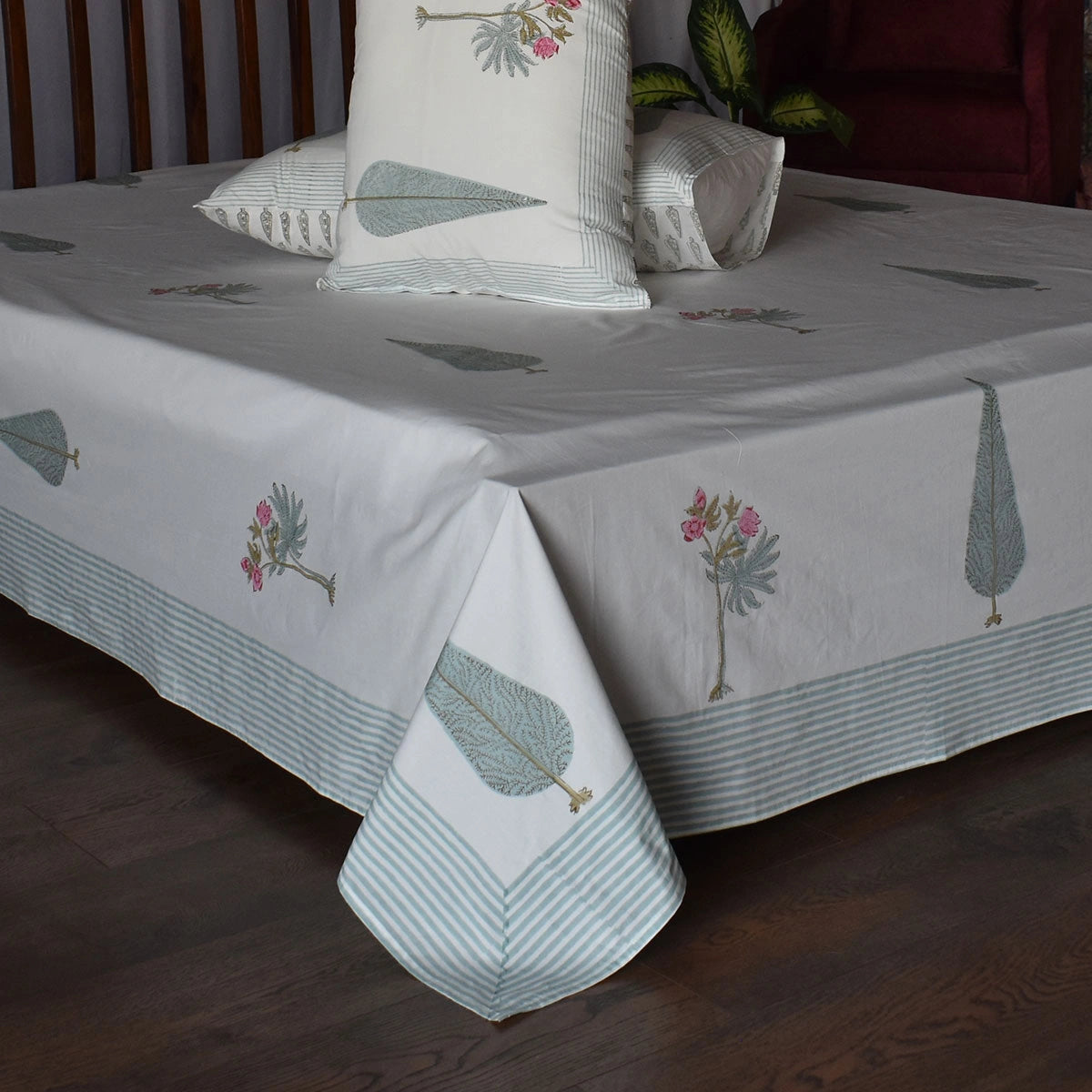 A bed dressed with a white tablecloth featuring a light blue striped border and scattered botanical leaf prints in muted blue-gray and pink, styled with matching pillows in a bedroom setting.