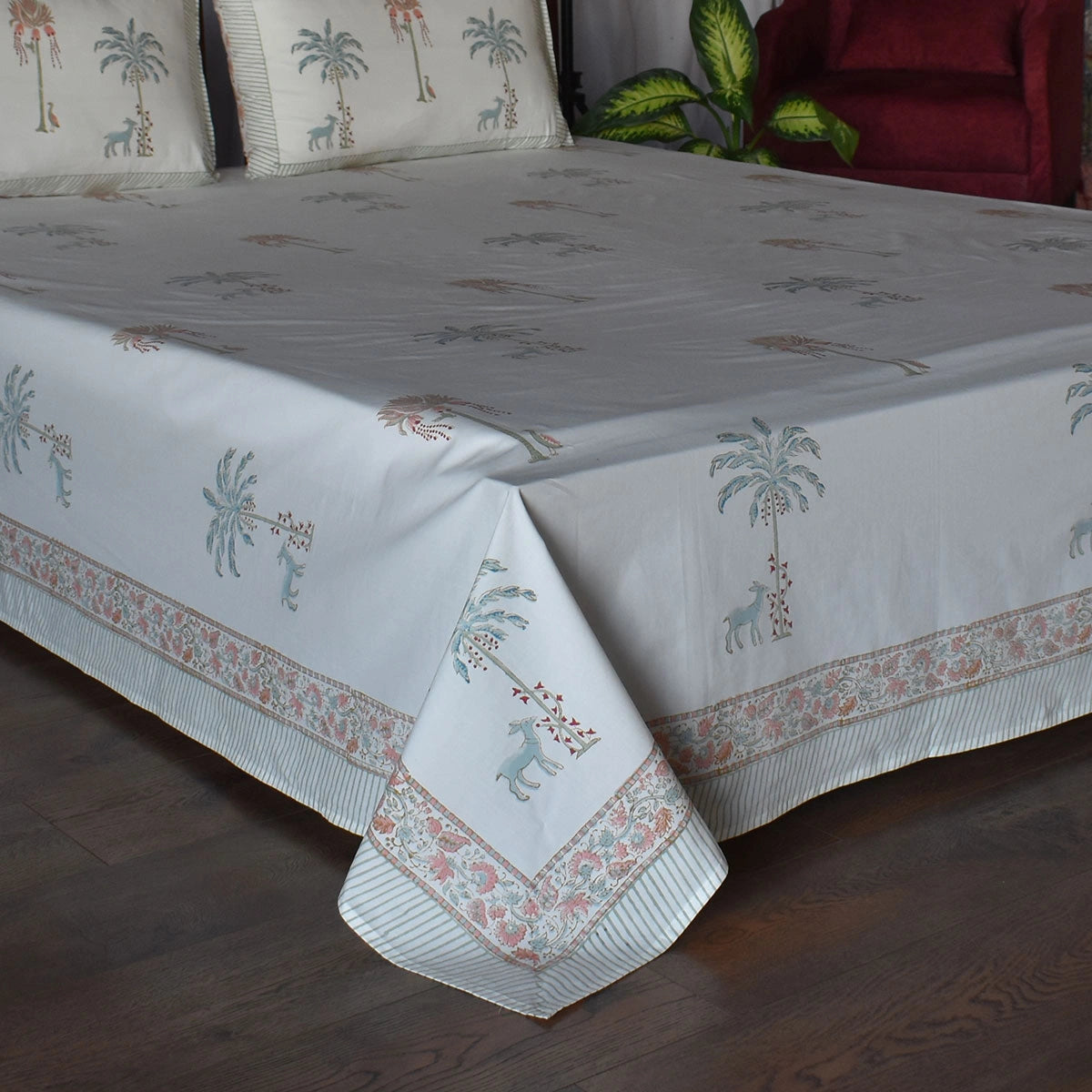A white cotton bedspread with a tropical print of palm trees, giraffes, and zebras, styled on a bed with matching pillowcases in a bedroom setting.