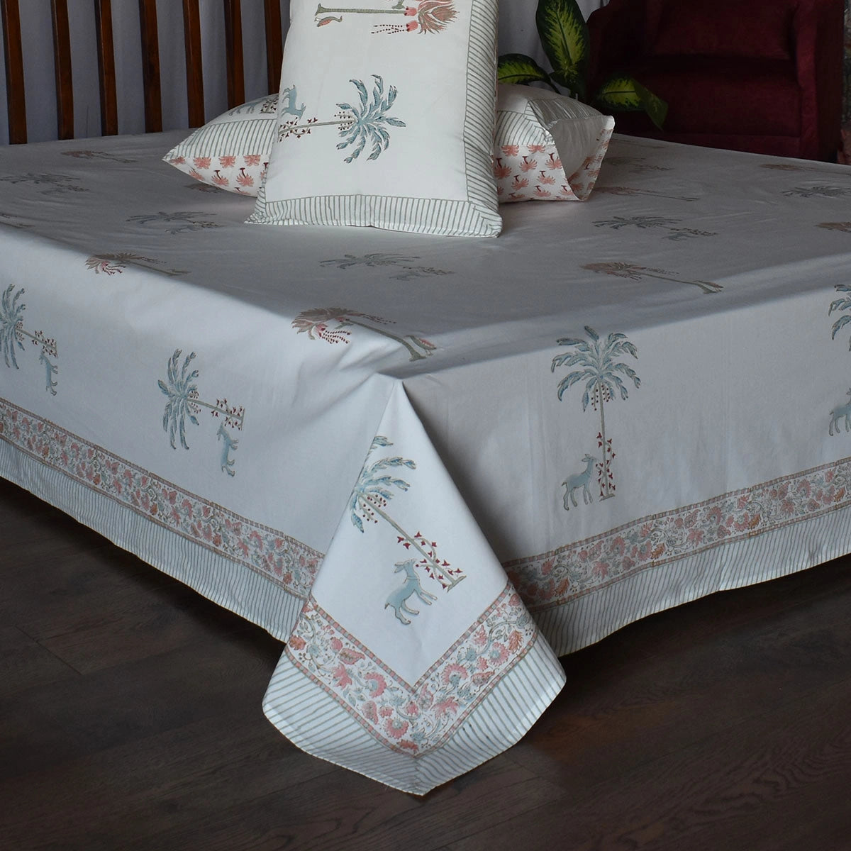 A white cotton bedspread set with a tropical print featuring palm trees, giraffes, and a decorative border, styled on a bed with matching pillows.