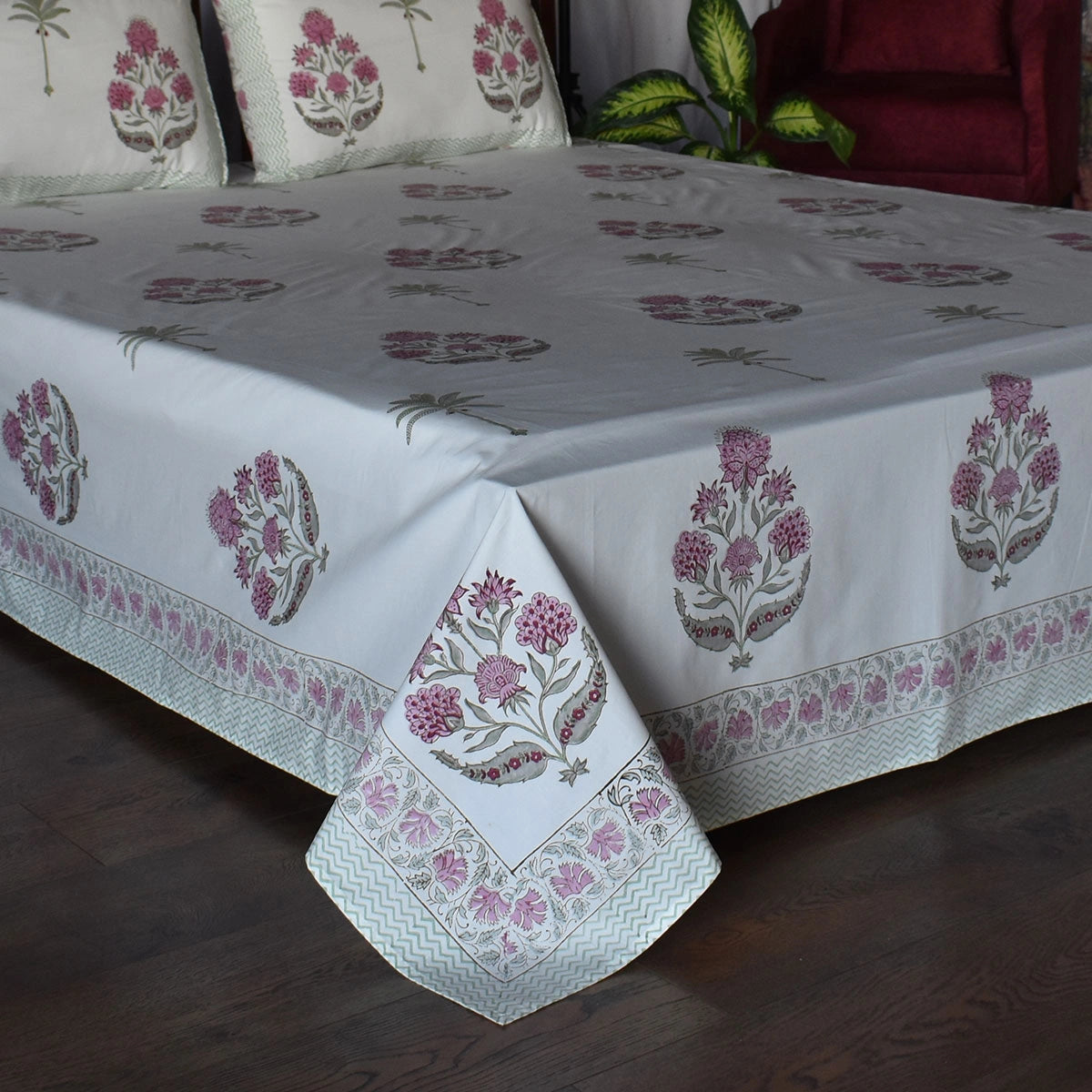 A white cotton bed sheet with pink and gray block-printed floral motifs and a decorative border, shown on a bed in a bedroom setting.