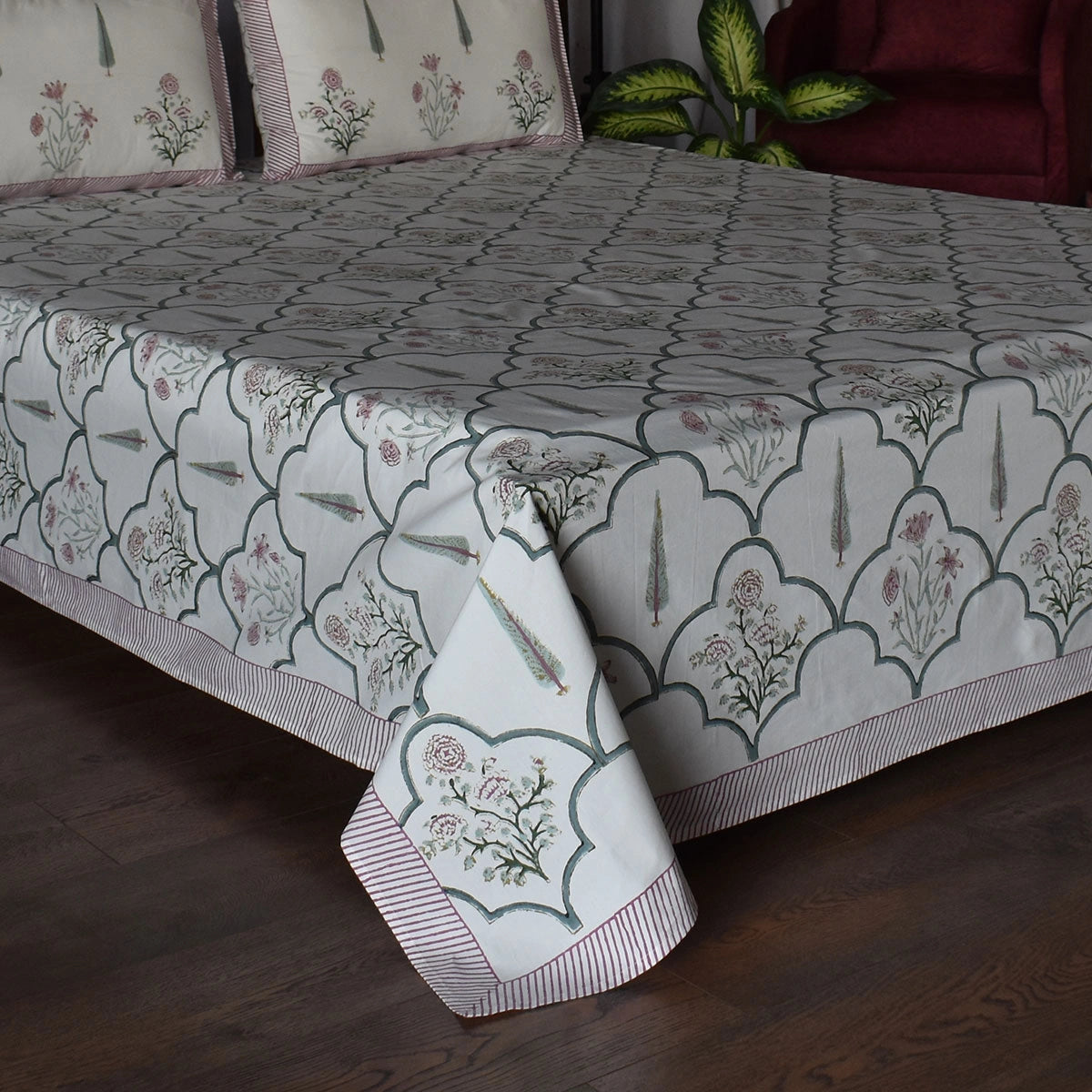 A patterned cotton bedspread and matching pillowcases on a bed, featuring a gray and white scalloped geometric design with pink floral motifs and striped borders, set in a bedroom with a dark wood floor and a red armchair in the background.