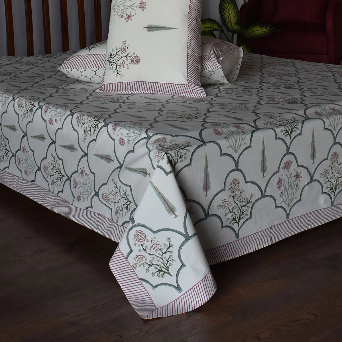 A patterned bedspread and matching pillows on a bed in a bedroom setting, featuring a white base with gray damask-style arch motifs, pink floral details, and striped borders.
