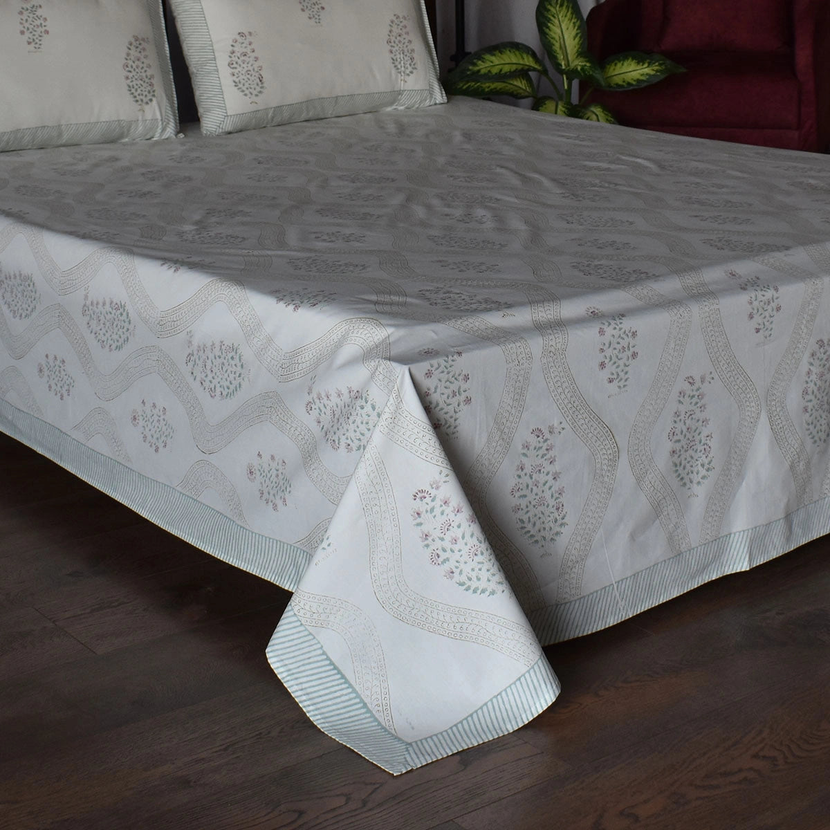 A light gray and white patterned bedspread with matching pillowcases on a bed, set against a dark wood floor with a red armchair and green plant in the background.