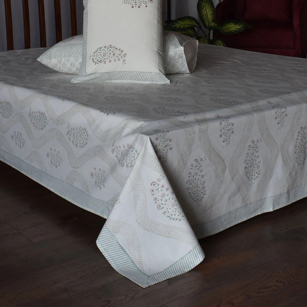 A white and light gray patterned bed sheet and matching pillowcases are draped over a bed in a bedroom setting with dark wood flooring and a red armchair visible in the background.