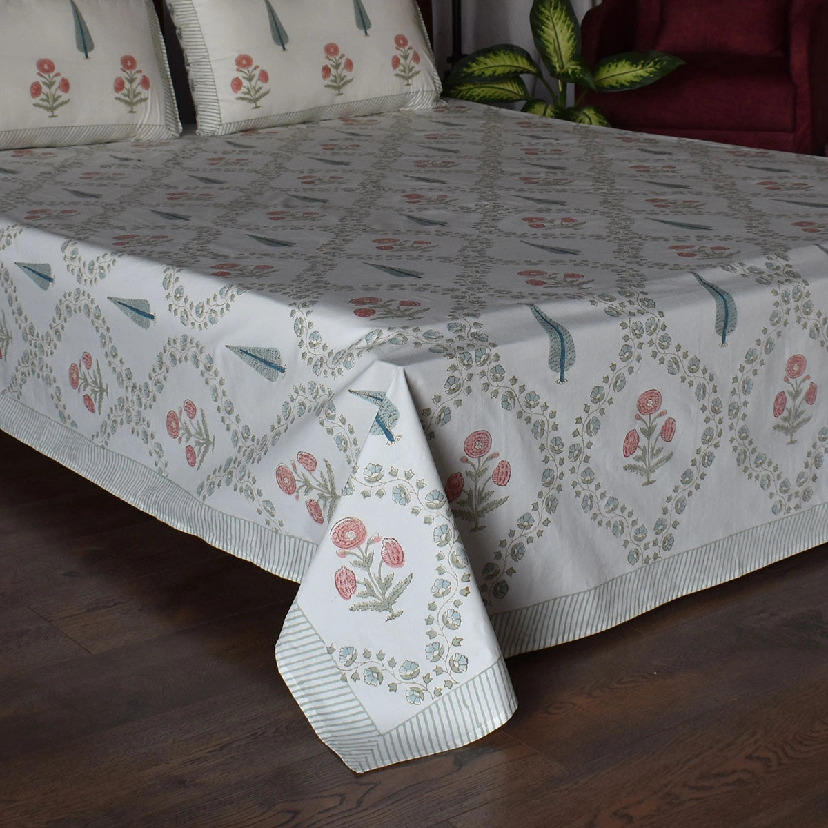 A floral printed cotton bed sheet with matching pillowcases on a bed, featuring a white base with pink and green botanical motifs and striped borders, set in a bedroom with a wooden floor and a plant in the background.