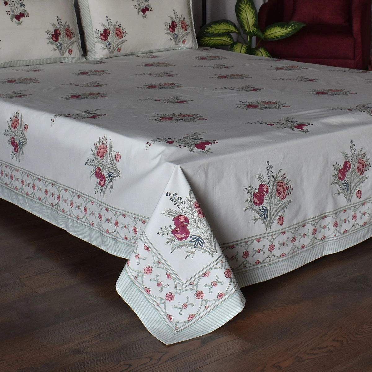 A white cotton bedspread with a delicate pink and green floral pattern and a matching striped border, draped over a bed in a bedroom setting with a dark wood floor and a red armchair in the background.