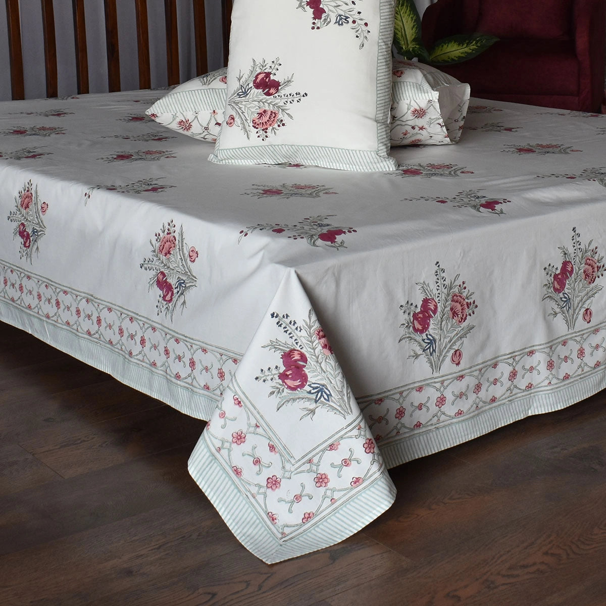 A white tablecloth with a pink and green floral pattern and striped border, styled on a bed with matching pillows in a bedroom setting.