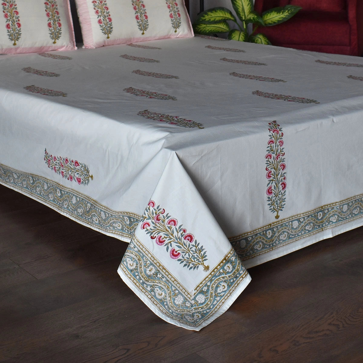 A white cotton tablecloth with traditional Indian block prints and borders, draped over a bed in a living room setting with a dark wood floor.
