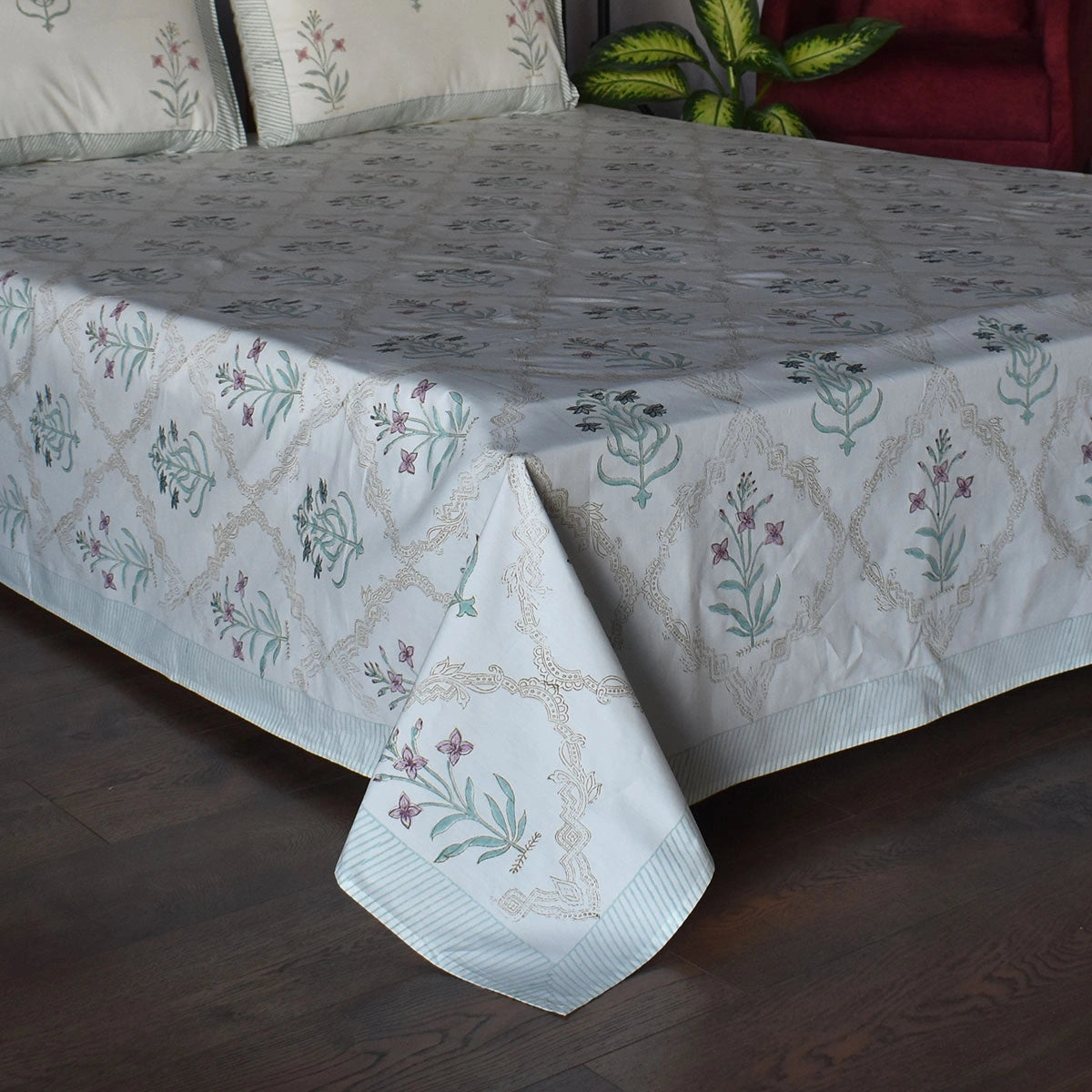 A light blue and white patterned bedspread with matching pillows and a decorative border, draped over a bed on a dark wood floor with a red sofa and green plant in the background.