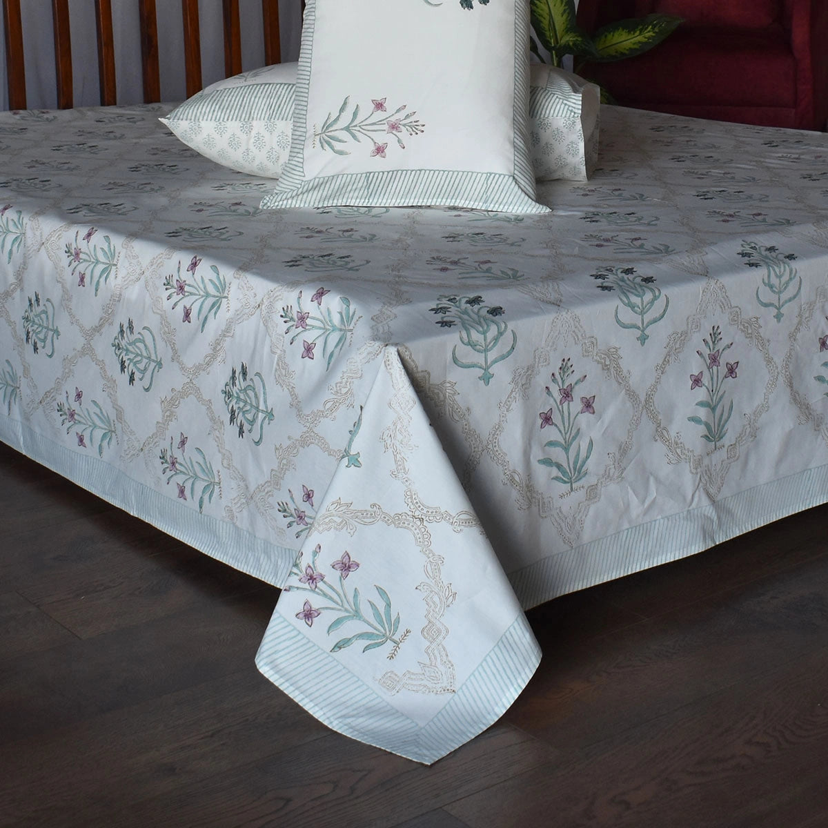 A white cotton bedspread and matching pillowcases featuring a delicate embroidered pattern of green vines, purple flowers, and beige trellis motifs, styled on a bed with dark wood flooring.
