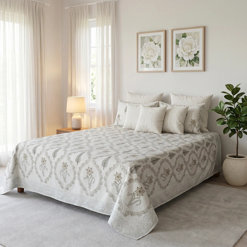 A neatly made bed in a softly lit bedroom, covered with a light beige and white damask-patterned quilt and matching shams. The room includes sheer curtains, a bedside lamp, framed floral wall art, a potted plant, and a light rug.