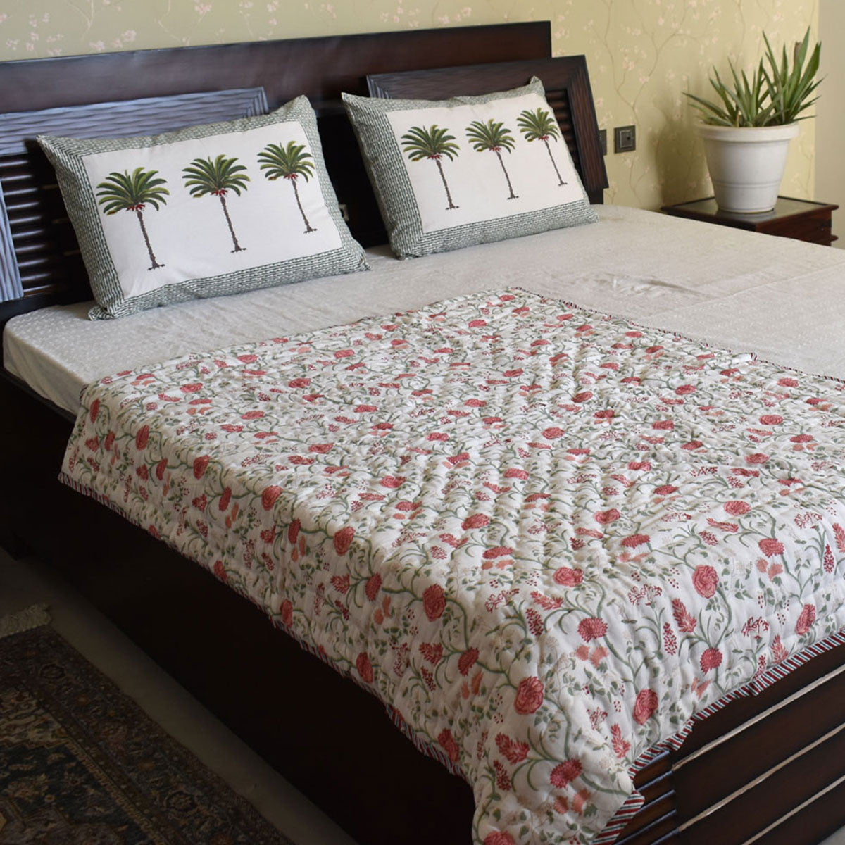 A quilted bedspread with a red and green floral pattern is spread across a dark wooden bed, accompanied by two matching pillow shams featuring a palm tree motif on a white background.
