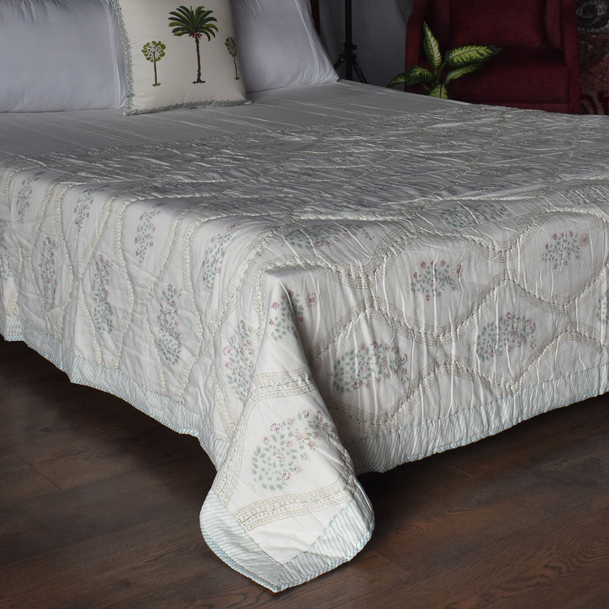 A quilted bedspread with a diamond stitched pattern and delicate floral embroidery in muted gray and soft pink, styled on a bed with matching pillows in a bedroom setting.