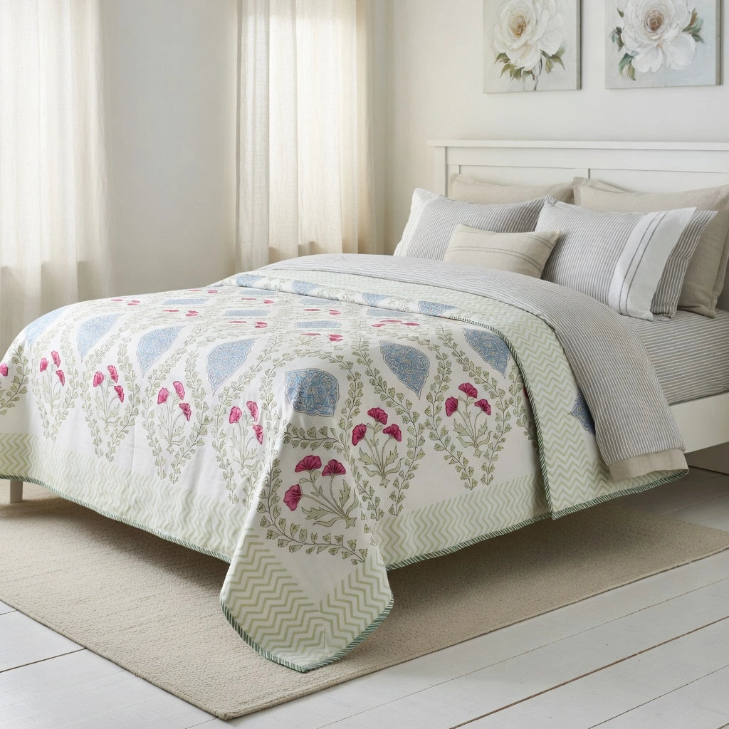 A quilted comforter set on a bed in a bright, neutral bedroom. The comforter features a cream base with a botanical pattern in sage green, light blue, and pink, layered over a striped gray and white sheet and pillows.