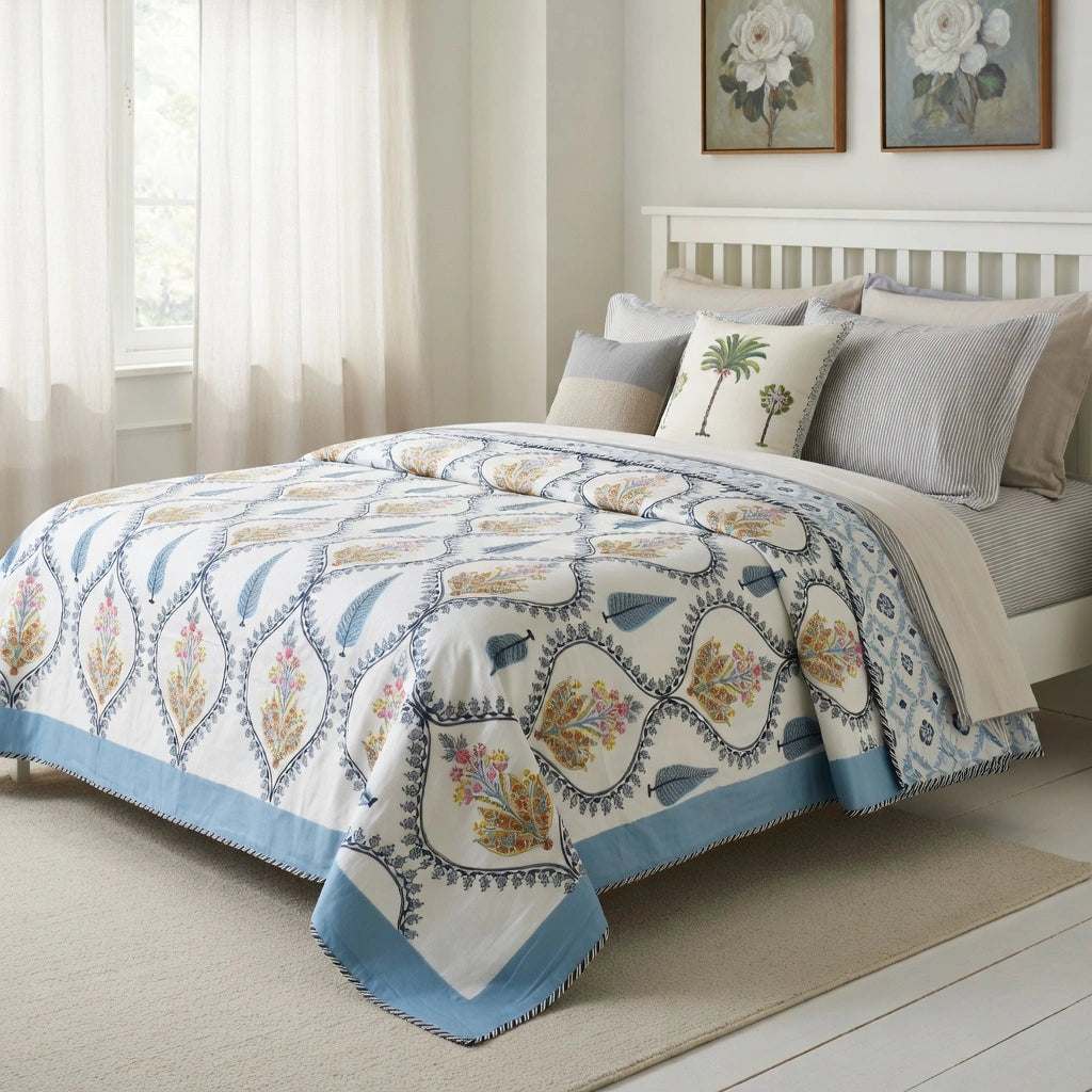 A patterned quilted comforter set on a bed in a bright bedroom. The comforter has a white base with a blue geometric trellis pattern and multicolored floral motifs, styled with gray and white pillows.