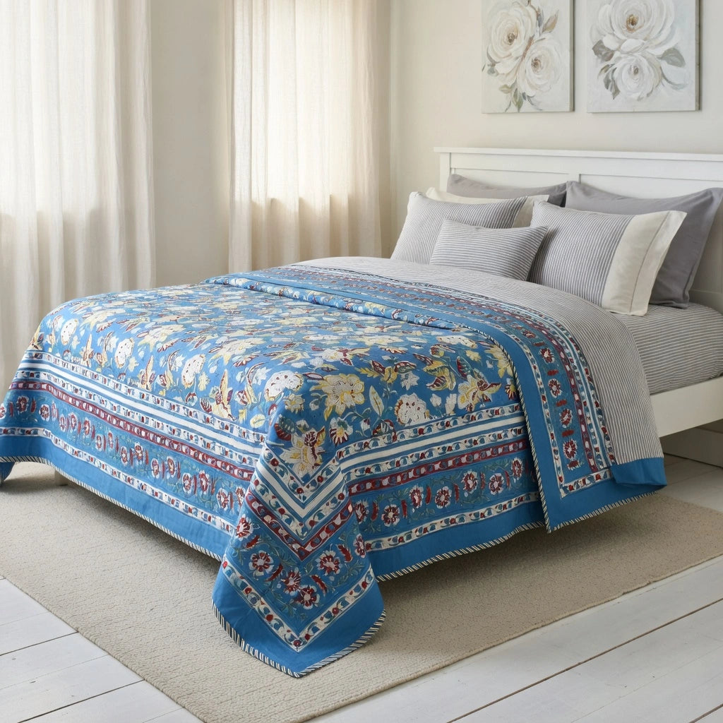 A blue patterned quilted comforter with intricate floral and geometric borders is styled on a bed in a bright, modern bedroom with white walls, sheer curtains, and neutral gray and white bedding.