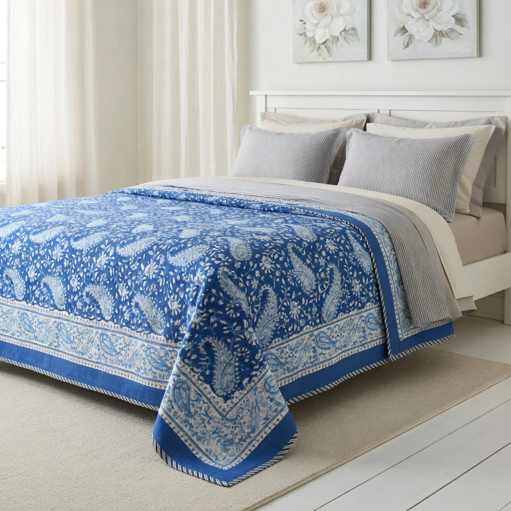 A blue and white paisley patterned quilted comforter set on a bed with gray pillows in a bright, neutral bedroom.