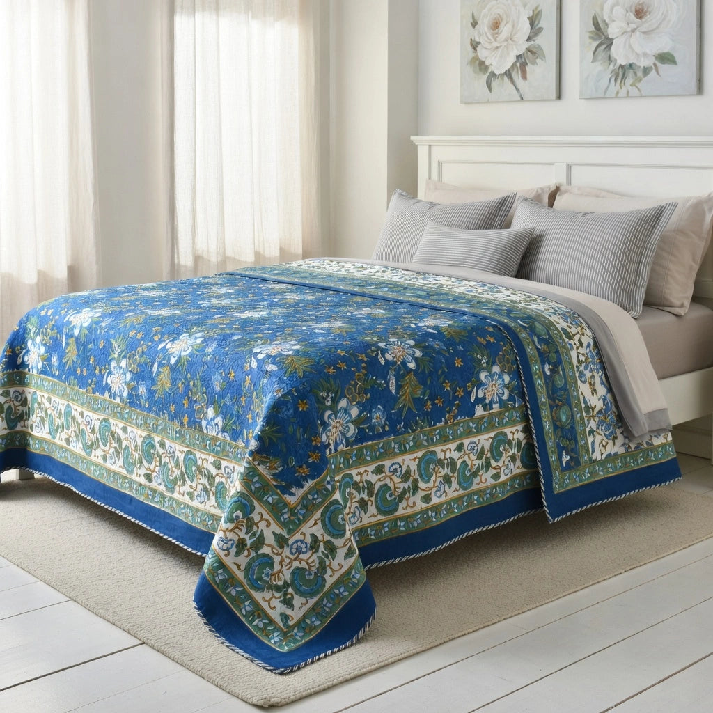 A blue and white floral patterned quilted comforter set on a bed in a bright, neutral bedroom. The comforter features a rich blue base with intricate white floral motifs and a decorative border in teal and cream.