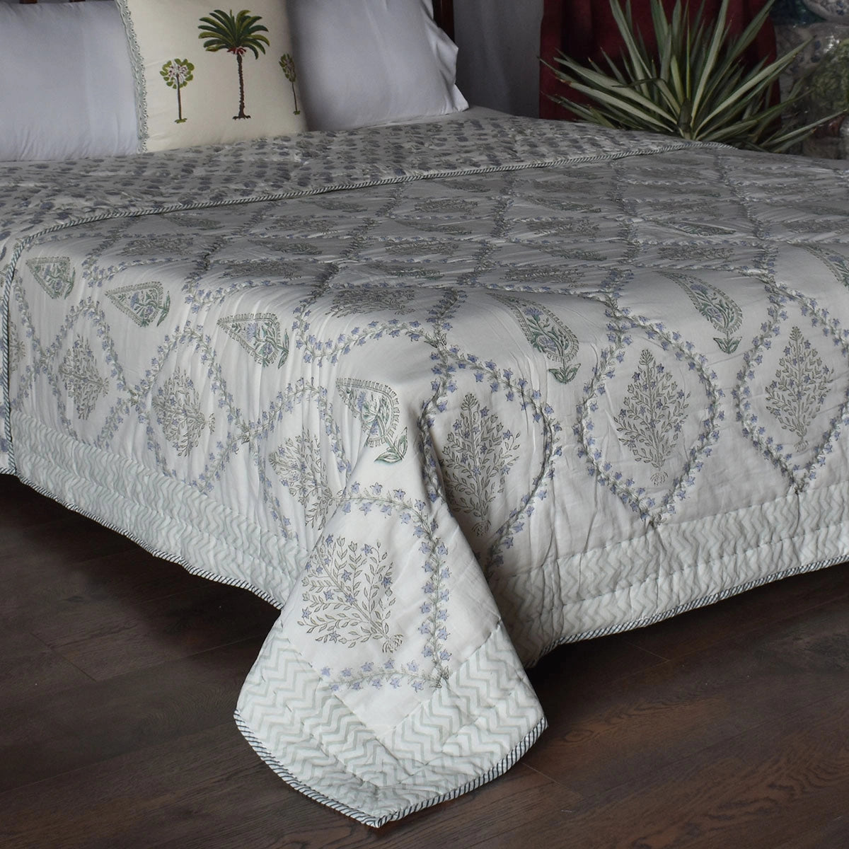 An embroidered quilted bedspread with a geometric and paisley pattern in gray and muted blue, styled on a bed with white pillows in a bedroom setting.