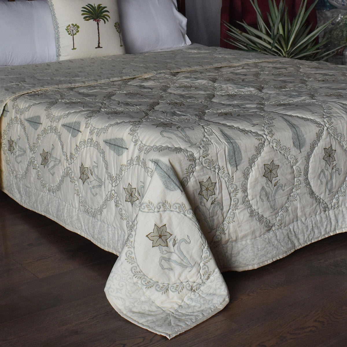 An embroidered quilted bedspread with a geometric and floral pattern in muted gray and beige tones, styled on a bed with matching pillows and a dark wood floor.