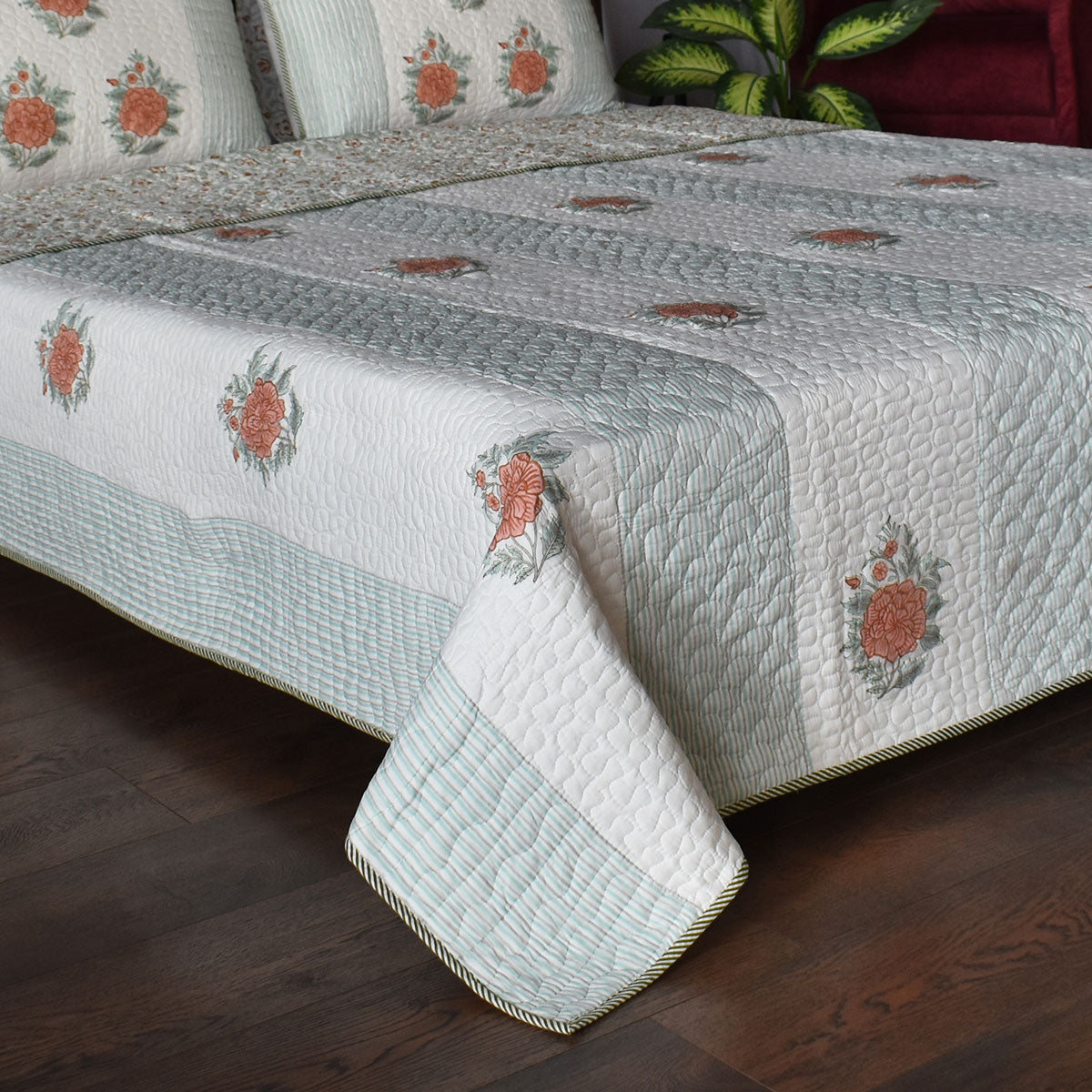 A quilted bedspread with matching pillow shams, featuring a white and pale blue-green background with orange floral motifs and a striped border, styled on a bed over a dark wood floor.