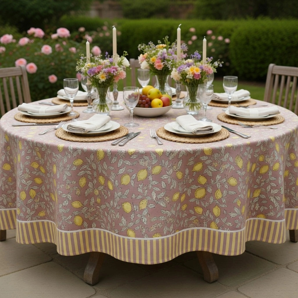 A round outdoor dining table covered with a dusty pink tablecloth printed with yellow lemons and green leaves, set with place settings, floral centerpieces, and candles in a garden setting.