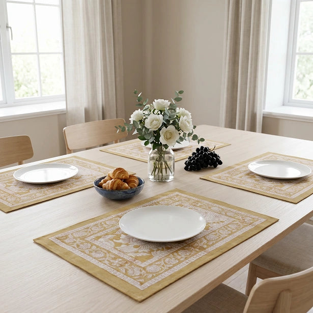 Four rectangular table placemats with a beige and white floral pattern arranged on a light wood dining table. The setting includes white plates, a glass vase with white flowers, a bowl of pastries, and a bunch of dark grapes, with chairs and windows in the background.