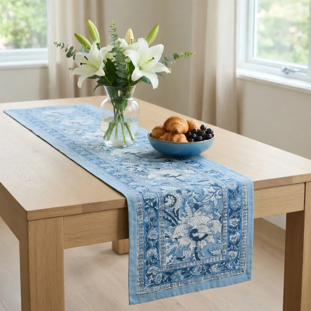 A blue and white patterned table runner draped over a light wood dining table, styled with a glass vase of white lilies and a blue bowl of pastries and blueberries in a bright room.