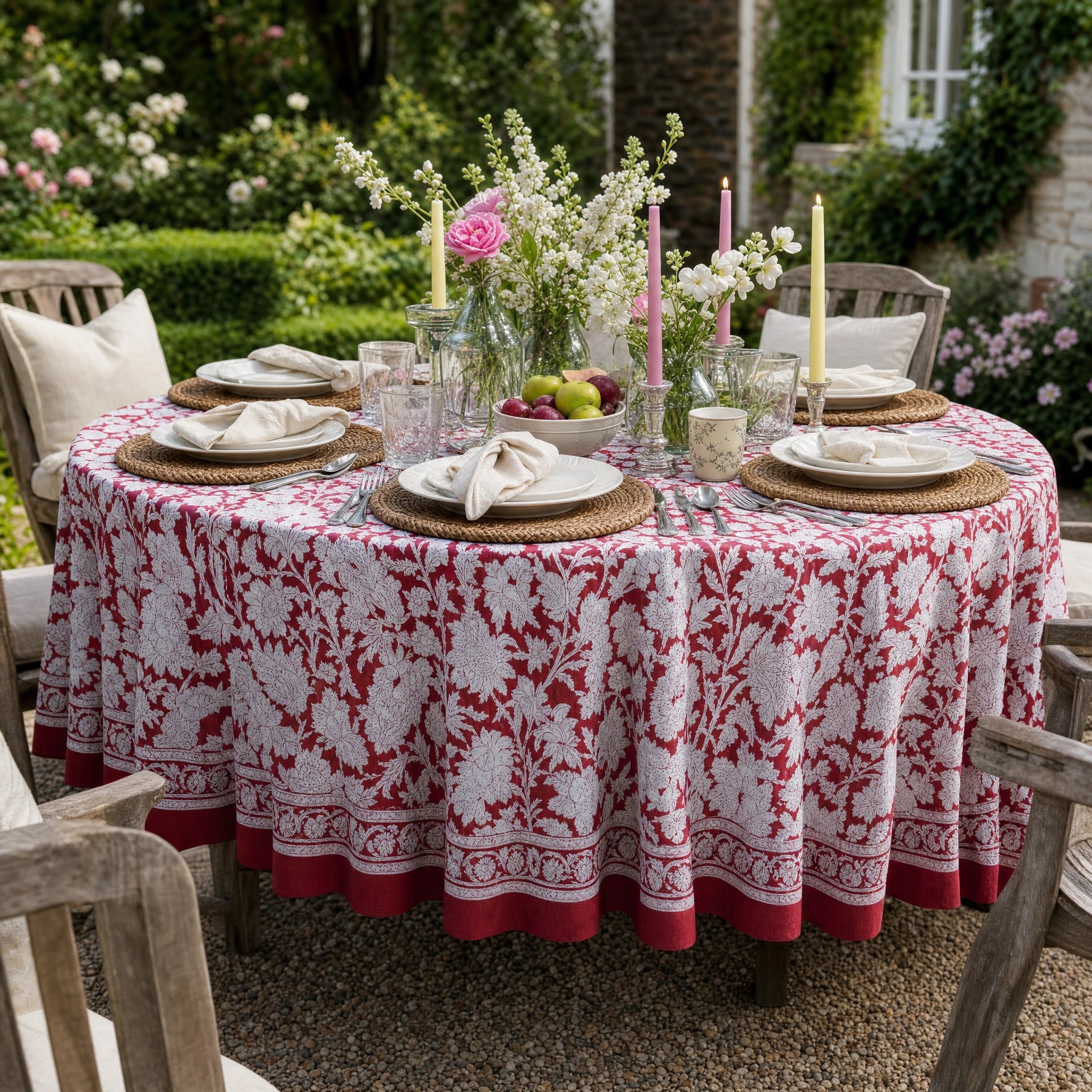 A round outdoor dining table covered with a red and white floral tablecloth, set with plates, folded napkins, woven placemats, glassware, and floral centerpieces in a garden setting.