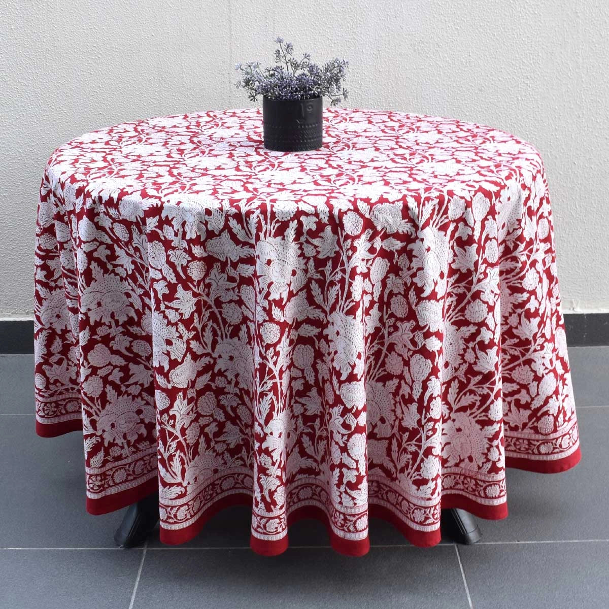 A round table covered with a red and white floral patterned tablecloth, with a small black planter on top, set against a light gray wall and dark gray tiled floor.