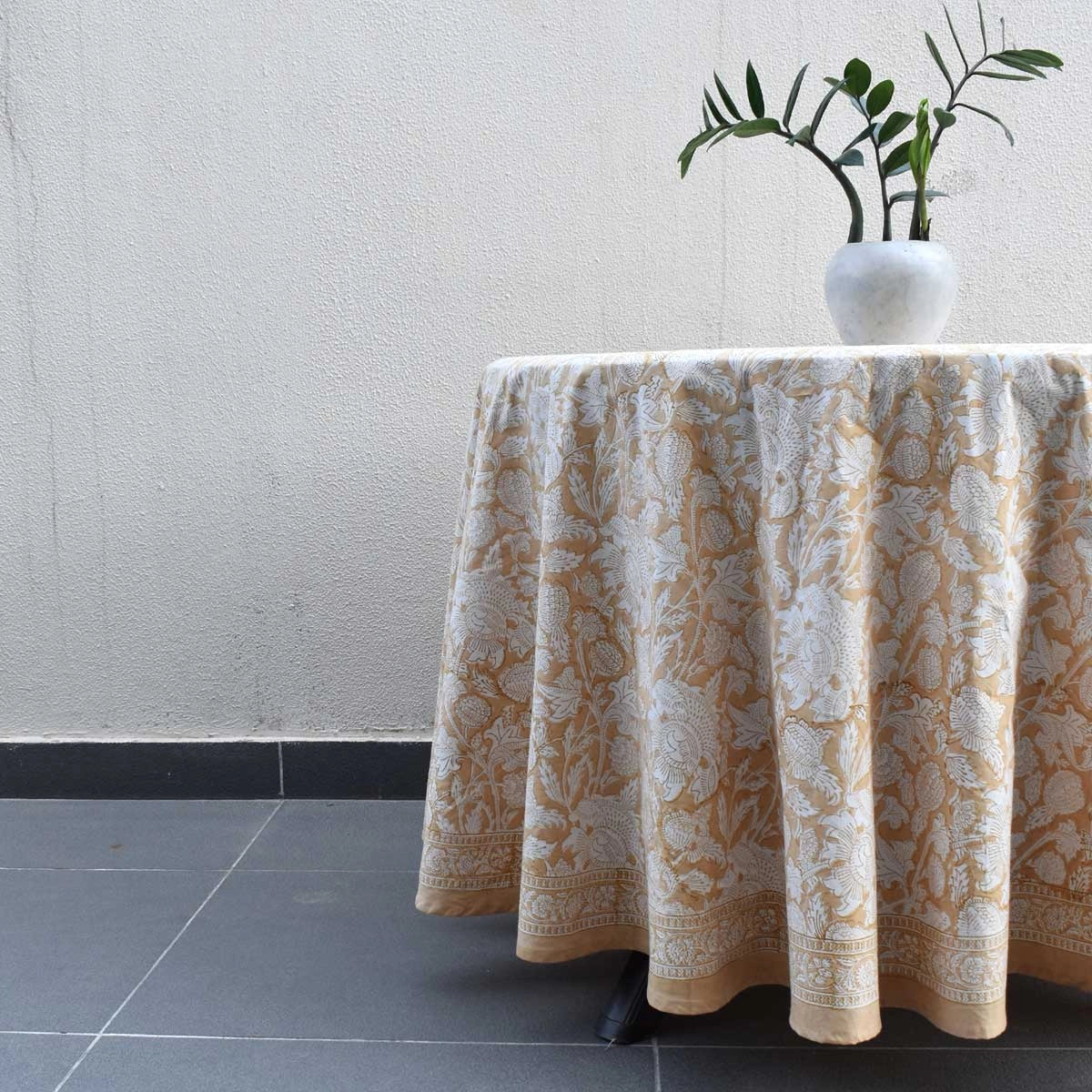 A round table draped in a beige and white damask patterned tablecloth, with a white vase containing green leaves on top, set against a textured off-white wall and dark gray tiled floor.