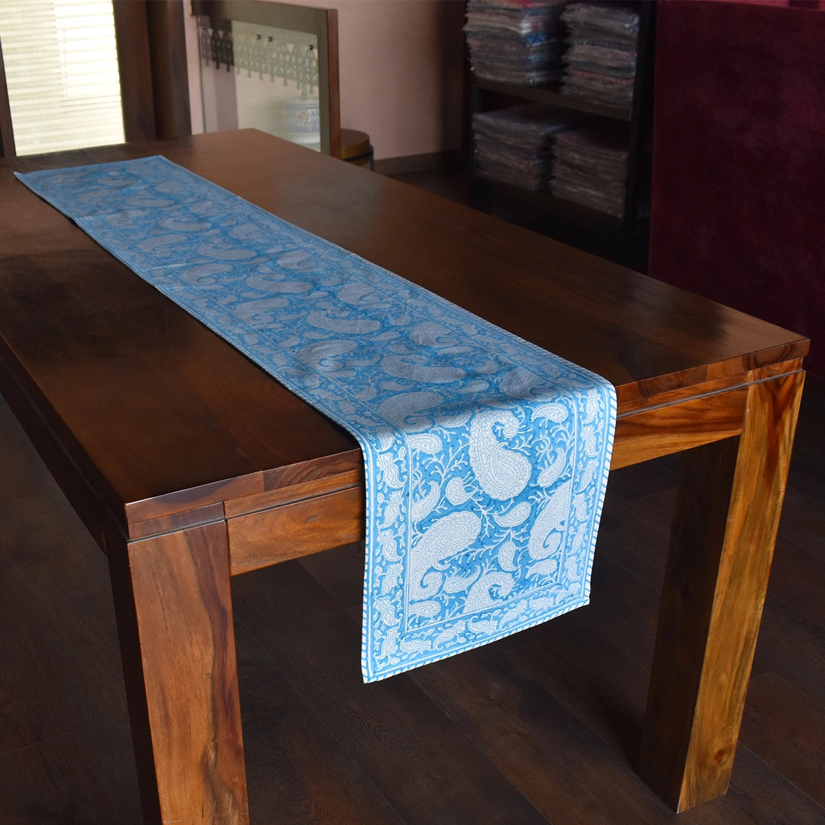 A light blue table runner with a white paisley pattern draped over a dark brown wooden dining table in a home setting.