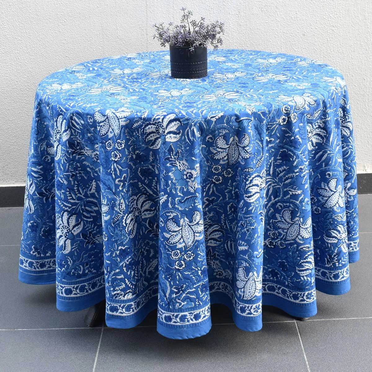 A round blue tablecloth with a white floral and butterfly pattern, draped over a round table with a small black vase of dried flowers on top, set against a light gray wall and dark gray tiled floor.