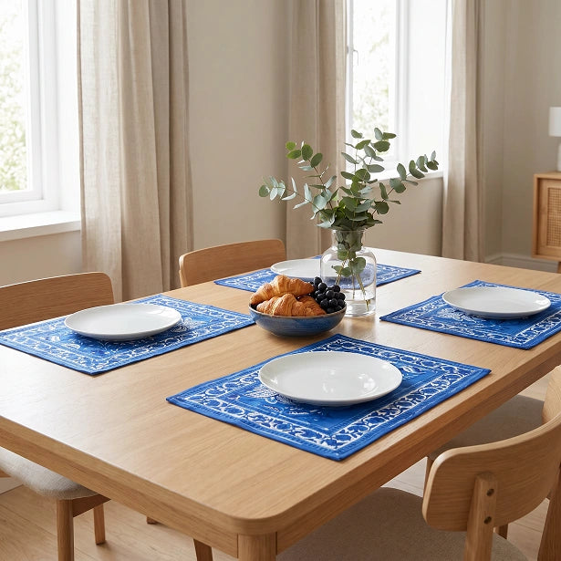 A dining table set with four blue placemats featuring a white ornamental pattern, white plates, a bowl of pastries, and a vase with greenery in a bright, modern dining room.