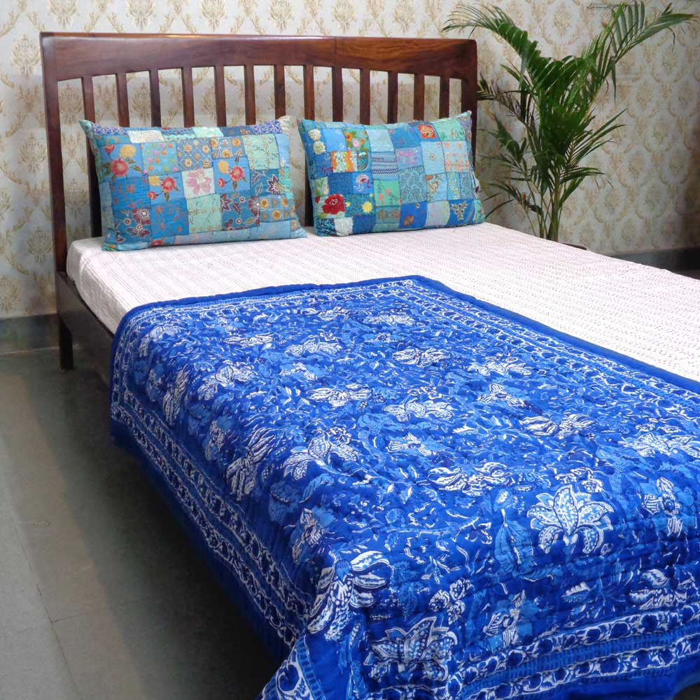 A blue and white patterned quilted bedspread with matching patchwork pillows on a wooden bed frame, set against a patterned wallpaper background.