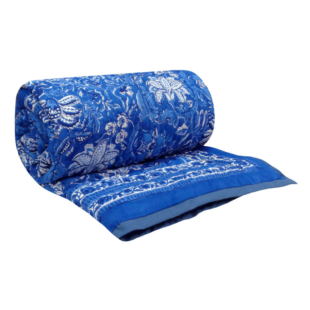 A blue quilted comforter with a white floral and paisley bandhani pattern, shown rolled up with one corner folded over on a plain white background.