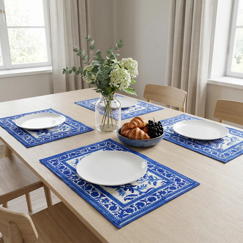 Blue and white patterned placemats set on a light wood dining table, styled with white plates, a glass vase of flowers, and a bowl of pastries in a bright dining room.