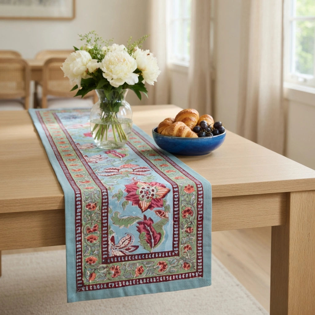 A light blue embroidered table runner with red, green, and white floral patterns is styled on a light wood dining table, flanked by a vase of white flowers and a blue bowl of pastries.