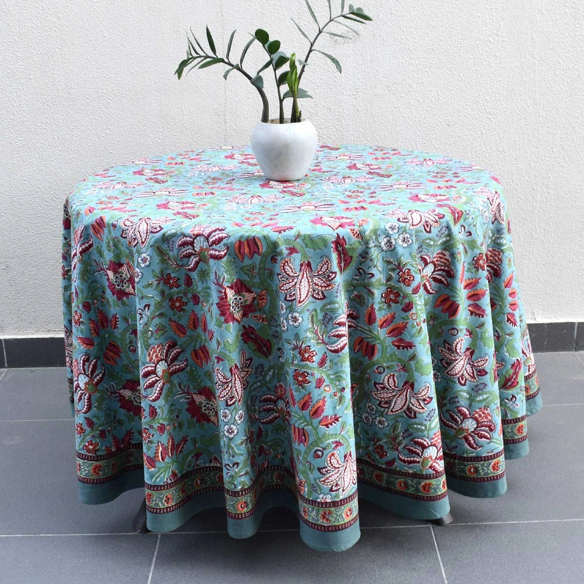 A round table covered with a light blue tablecloth featuring a red and pink floral pattern, with a white vase containing green leaves on top, set against a white wall and gray tiled floor.