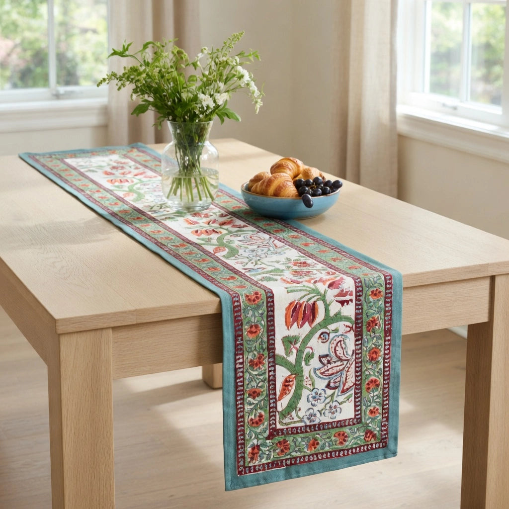 A rectangular table runner with a detailed floral and butterfly pattern in teal, red, green, and cream, styled on a light wood dining table with a vase of flowers and a bowl of fruit.