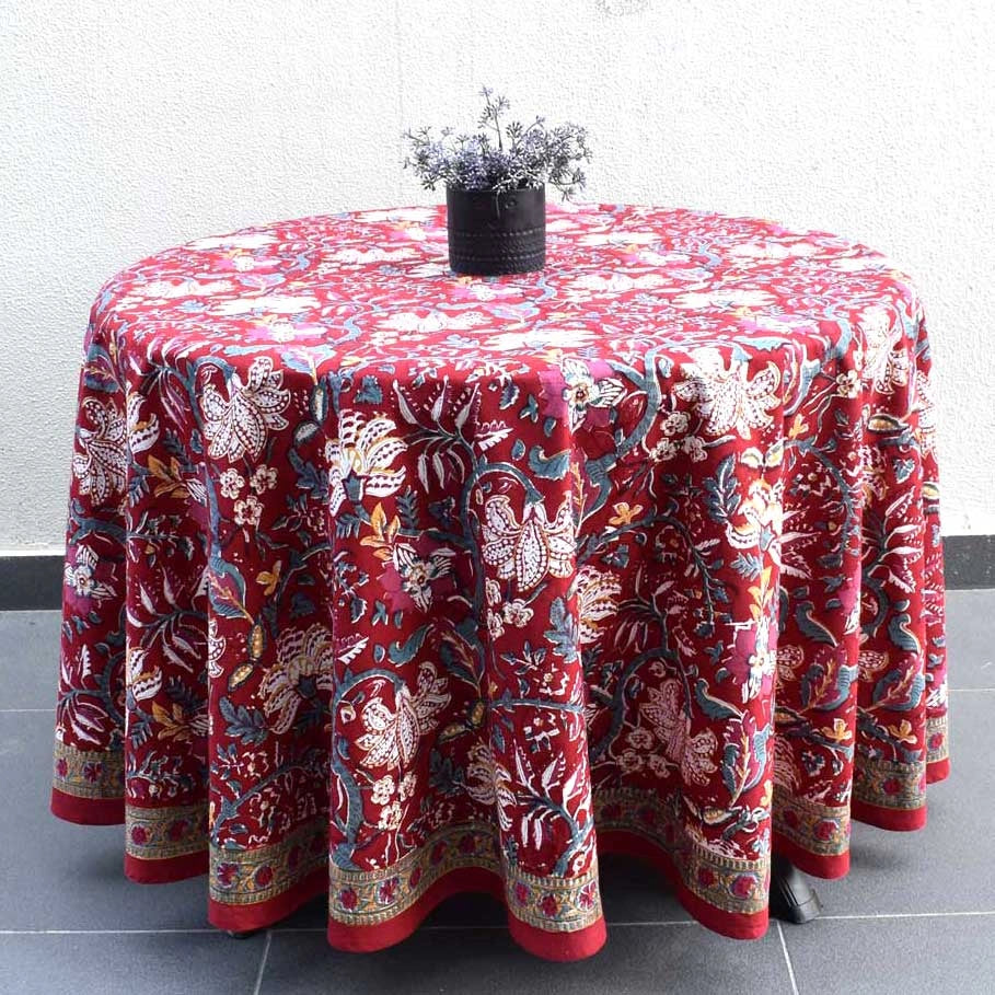 A round table covered with a richly patterned red tablecloth featuring intricate floral and bird motifs in cream, blue, and gold, styled with a small black vase of dried lavender on top against a white wall.